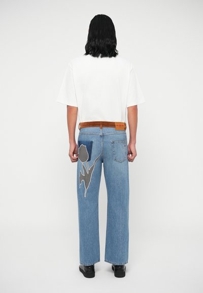 Világoskék farmer széles lábú nadrág, hátul szürke virágmintával hímzett zsebbel, sima fehér pólóval és barna övvel párosítva.