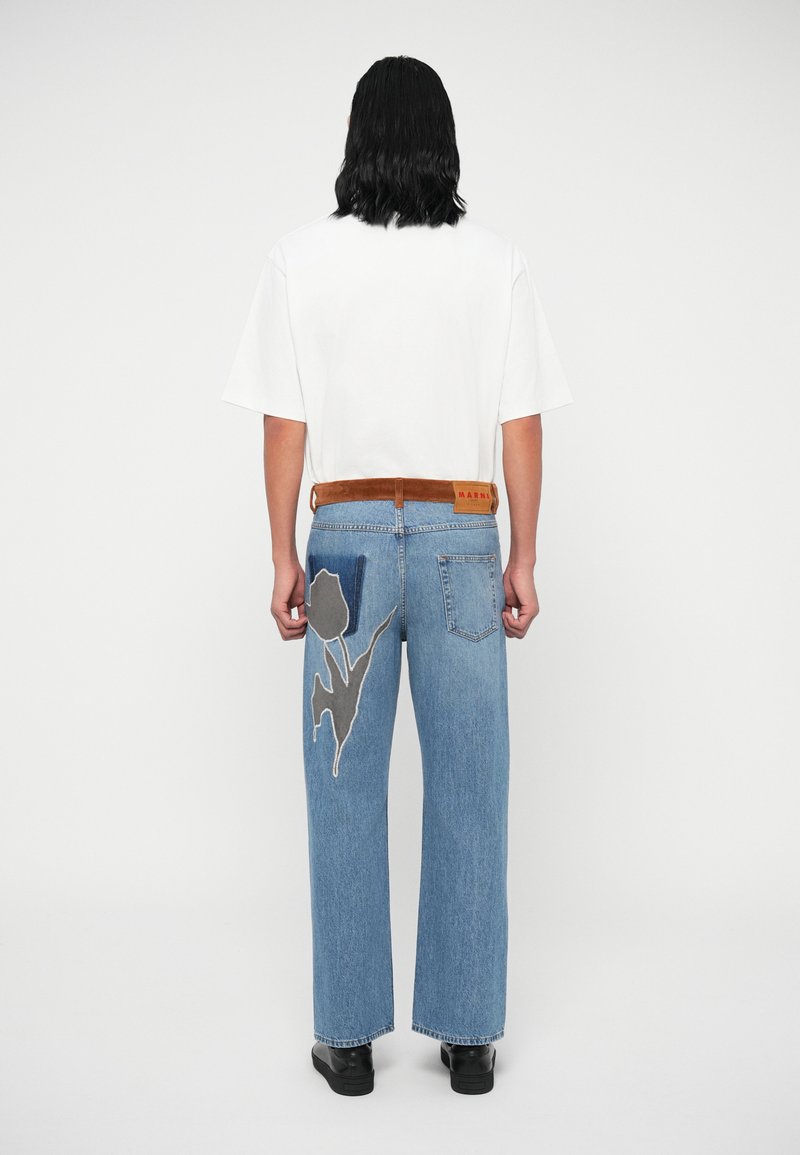 Helle blaue Jeans mit weitem Bein, die mit einer grauen Blumenstickerei auf der hinteren Tasche verziert sind, kombiniert mit einem schlichten weißen T-Shirt und einem braunen Gürtel.