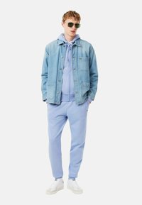 Veste en denim bleu clair, à manches longues, avec fermeture à boutons. Superposée sur un hoodie bleu clair, associée à un pantalon de jogging assorti et des baskets blanches.