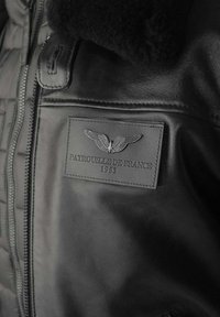 Veste en cuir noir avec col en fourrure, dotée d'un patch texturé embossé avec "PATROUILLE DE FRANCE 1953" et un design d'aile.