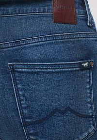 Granatowe dżinsy z ciemnego denimu z brązową skórzaną metką z napisem "Mustang" na pasku oraz małym logo na tylnej kieszeni.