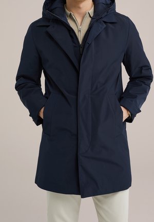 Homme portant un manteau à capuche bleu marine, les mains dans les poches, par-dessus un pull beige zippé et un pantalon de couleur claire.
