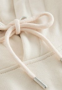 Gros plan d'un cordon en tissu crème noué en nœud avec des embouts métalliques et des détails cousus sur un matériau doux.