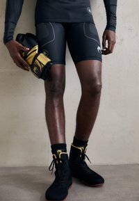 Pantaloni sportivi neri con cuciture a contrasto, abbinati a scarpe alte nere con accenti gialli. Viene tenuto un guantone da boxe imbottito nero e oro.