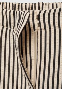 Gros plan sur un tissu beige à rayures noires verticales montrant une fermeture éclair ouverte et deux crochets métalliques sur la ceinture d'un vêtement.