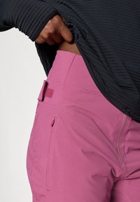 Roze technische broek met een gladde structuur, ritszakken en verstelbaar klittenband in de taille, gecombineerd met een donkere lange mouwen shirt.
