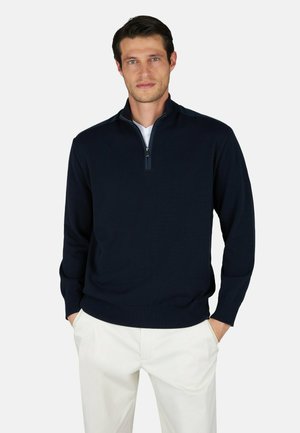 Homme portant un pull bleu marine à demi-fermeture éclair par-dessus une chemise blanche et un pantalon blanc, debout les mains dans les poches devant un fond uni.