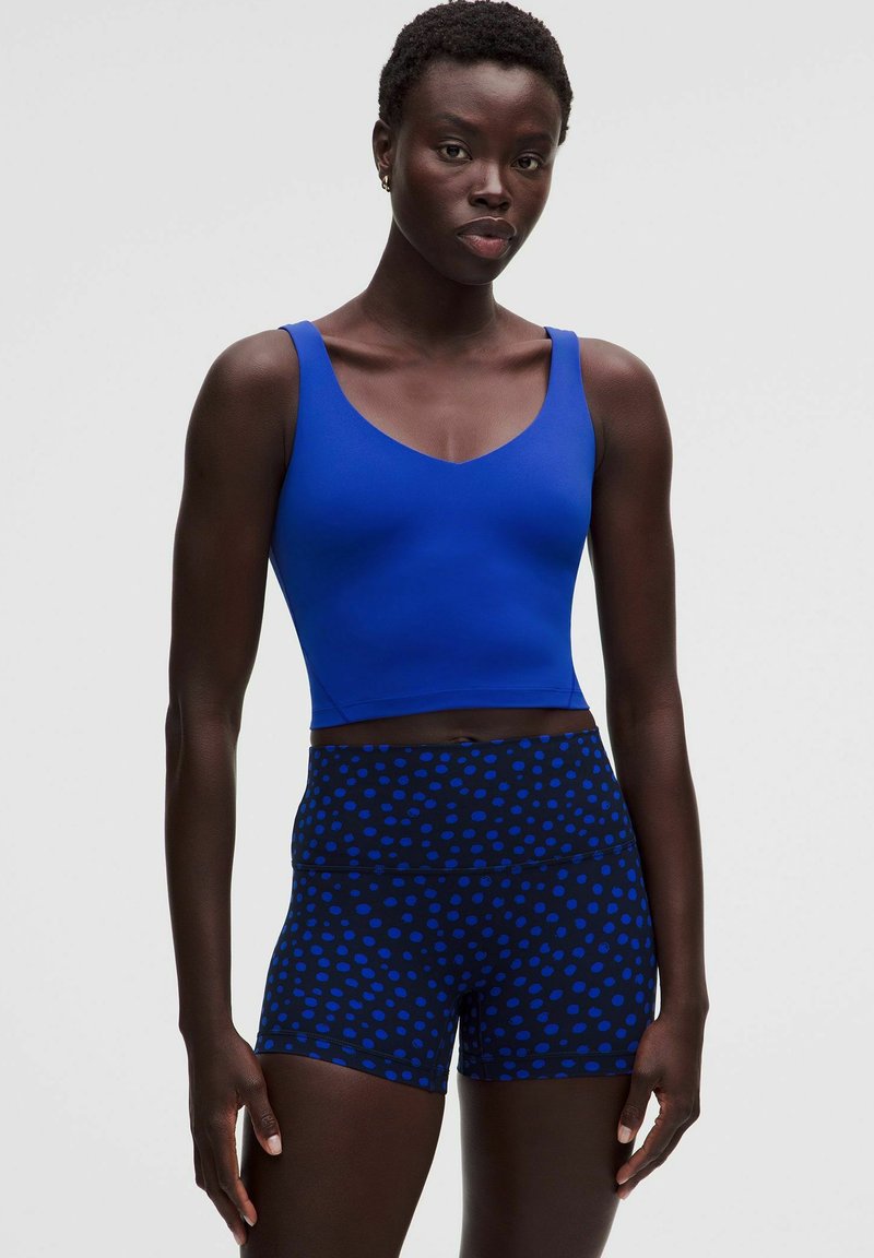 Woman wearing a blue sleeveless crop top and black shorts with blue polka dots, standing against a plain light background.