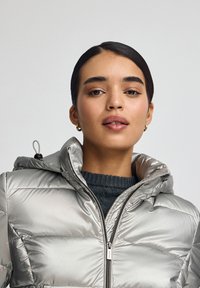 Silverfärgad vadderad jacka med huva, med quiltad textur och dragkedja framtill. Bärs över en mörkstickad tröja.