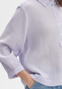 Camicia lavanda con colletto in tessuto testurizzato e a coste, con bottoni e maniche ampia, abbinata a jeans azzurri chiari.