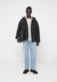 Sort pufferjakke med hætte, beige skjorte med knapper, lyseblå jeans og sorte sko, holder en sort lædertaske.