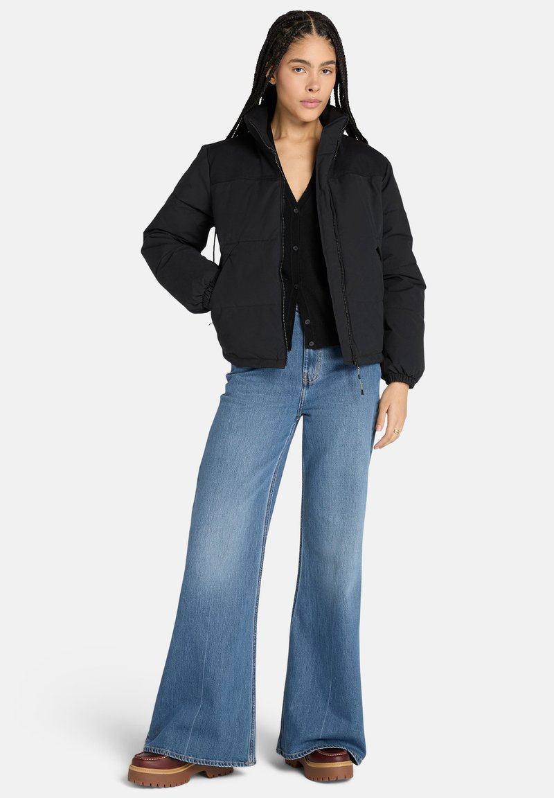 Giacca a vento nera con colletto alto, polsini elasticizzati e chiusura con zip, abbinata a jeans azzurri a gamba larga e stivali platform marroni.