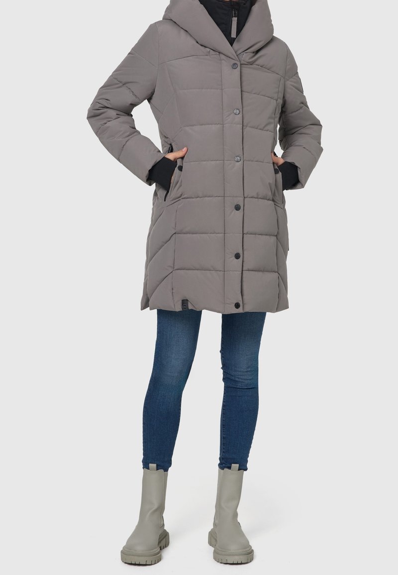 Grå pufferjakke med hætte, prydet med vandret quiltning, sort kant og knaplukning. Båret over blå jeans og grå ankelstøvler.