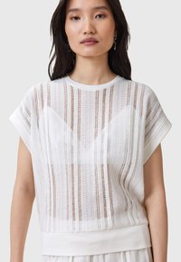 Witte doorschijnende top met horizontale strepen, zachte textuur, ronde hals en korte mouwen. Bevat een effen witte band aan de onderkant.