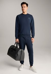 Sweatshirt blu navy e pantaloni jogger realizzati in tessuto morbido, abbinati a una borsa a tracolla nera testurizzata con un motivo geometrico e manici neri.