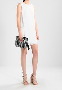 Bílé šaty bez rukávů, nad kolena. Černobílá taška ve tvaru chevron s třásněmi. Béžové sandály na vysokém podpatku se pásky. Hladký textilní materiál.