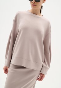 Ljus taupe sweatshirt med avslappnad passform, rund halsringning, långa ärmar och synliga sömnadsdetaljer, tillsammans med en matchande kjol.