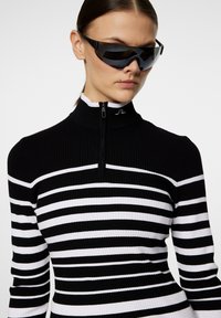 Svart och vitrandig ribbad långärmad topp med hög krage och halv dragkedja; kombinerad med eleganta svarta solglasögon.