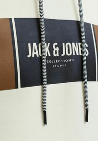 Felpa crema con stampa a blocchi marrone e blu. Presenta il logo "JACK & JONES" in bianco e lacci a zigzag in contrasto.