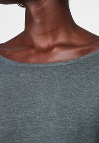 Harmaa neulepaita, jossa on leveä kaula-aukko, ja joka on varustettu sileällä pinnalla sekä röyhelöillä reunoissa. Lähikuva kankaasta ja ihonsävystä.