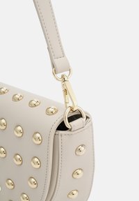 Sac à main en cuir beige avec des clous ronds dorés, doté d'une bandoulière amovible, de quincaillerie métallique et d'un détail de surface texturée.