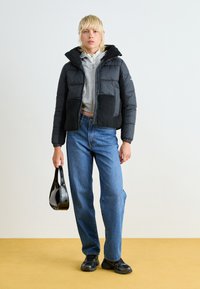 Svart dunjakke med en teksturert svart fleecepanel, grå hettegenser med glidelås, blå jeans og svart håndveske; modellen har på seg svarte sneakers.