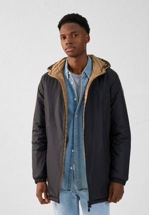 Veste isolée noire avec un intérieur matelassé beige, fermeture éclair et poignets réglables, superposée à une chemise en denim bleu clair.