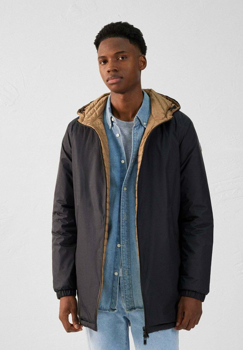 Chaqueta aislante negra con un forro acolchado beige, cierre con cremallera y puños ajustables, superpuesta a una camisa de mezclilla azul clara.