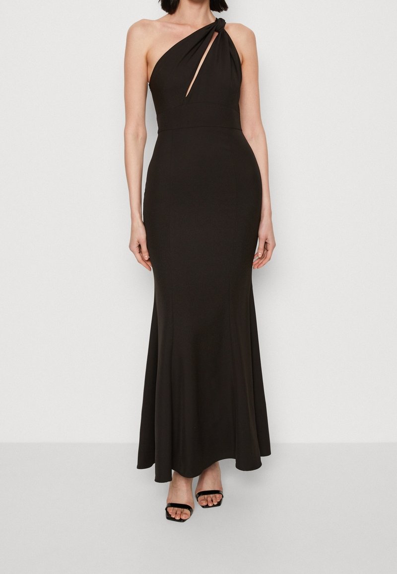 Femme portant une robe noire asymétrique avec une découpe avant et une jupe drapée, associée à des talons ouverts noirs.
