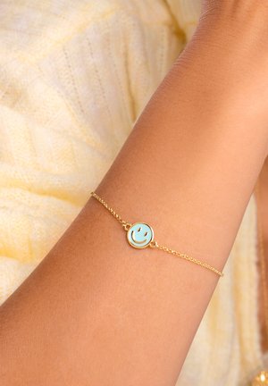 Goldene Kettenarmband mit einem runden Anhänger, der ein hellblaues Smiley-Gesicht zeigt. Die Kette ist zart mit einer feinen Textur und wird an einem gebräunten Handgelenk getragen.