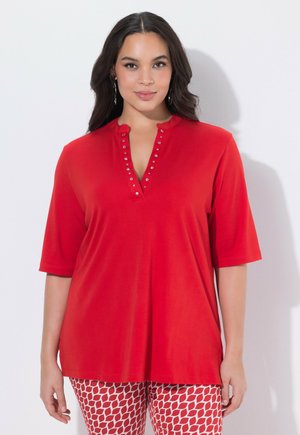 Donna che indossa una maglietta rossa a maniche corte con borchie argentate lungo la scollatura a V, abbinata a pantaloni rossi e bianchi con motivo a fantasia.