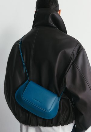 Persona che indossa una giacca di pelle nera con collo alto, con una piccola borsa a tracolla di pelle blu portata sulla schiena.