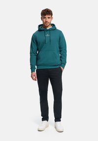 Sudadera con capucha color azul verdoso con bolsillo tipo canguro, capucha con cordón, puños y dobladillo acanalados. Combinada con pantalones oscuros y zapatillas blancas.