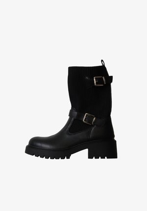 Botte noire mi-mollet avec semelle épaisse, dessus en tissu côtelé, deux boucles argentées réglables et une languette pour tirer en haut à l'arrière.