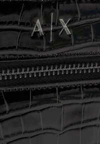 Närbild av svart läder med krokodilstruktur, med silvrig "A|X"-logotyp och synlig metallylla på en modeaccessoar.