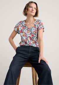 Šviesiai pilkas V-kaklo marškinėliai su gėlių raštu, akcentuota ryškiomis raudonomis, oranžinėmis ir mėlynomis spalvomis. Derinama su tamsiomis, laisvai krentančiomis džinsomis.