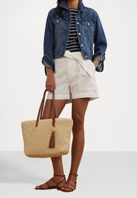 Woven straw tote bag with leather handles and tassel detail, paired with a denim jacket and striped T-shirt, and beige linen shorts.