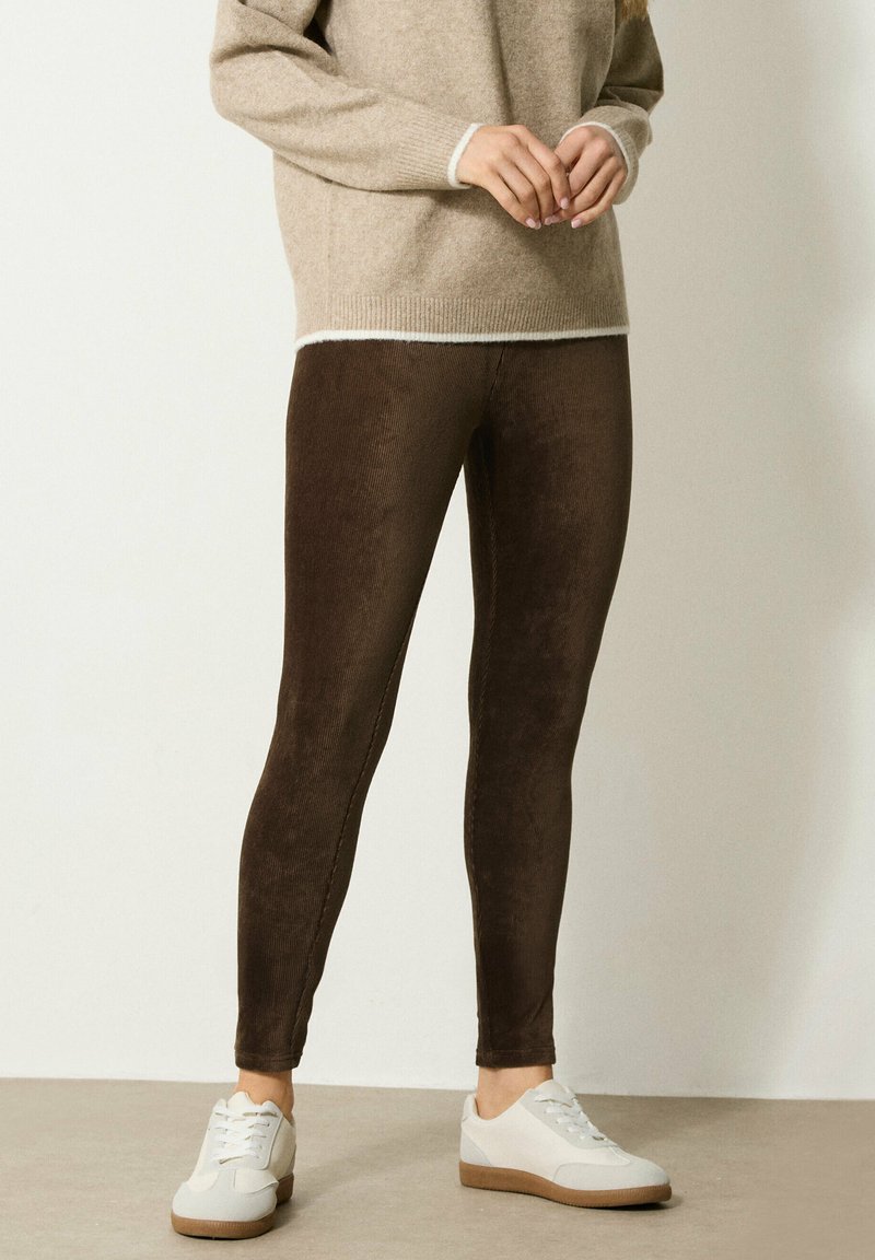 Vrouw in bruine corduroy legging, beige trui en witte sneakers, staand met handen voor zich gekruist tegen een effen achtergrond.