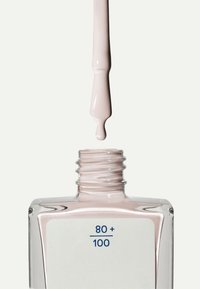 Botella de esmalte de uñas con base de vidrio transparente, líquido rosa pálido y una etiqueta blanca con texto azul. El pincel gotea esmalte sobre la abertura.