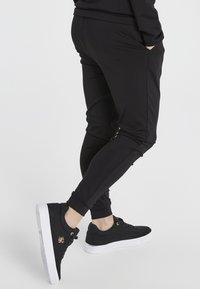 Schwarze sportliche Jogginghosen aus dehnbarem Material, mit seitlichen Taschen und schmaler Passform, kombiniert mit schwarzen Sneakers mit weißen Sohlen.
