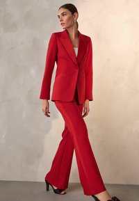 Punainen bleiseri lovikauluksilla ja yksirivisellä napituksella, yhdistetty samansävyisiin housuihin, joissa on sivuraidat, sekä mustiin korkokenkiin.