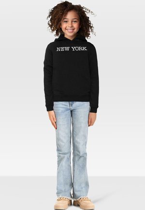 Smilende barn med krøllete hår iført svart hettegenser med "NEW YORK"-tekst, lyseblå jeans og beige klumpete sko som står på hvit bakgrunn.