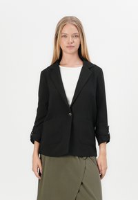 Zwarte blazer met één knoop, twee voorzakken en opgerolde mouwmanchetten. Draag het over een witte top en een olijfgroene, gedrapeerde rok.