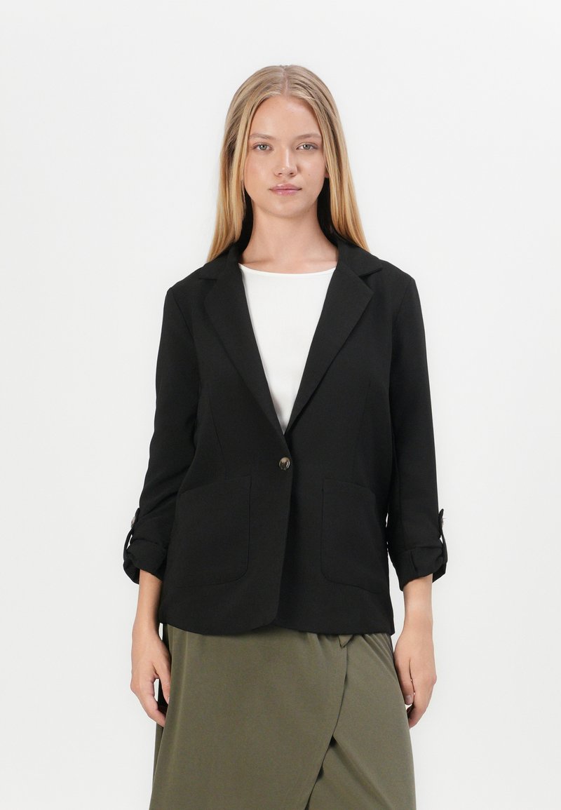 Zwarte blazer met één knoop, twee voorzakken en opgerolde mouwmanchetten. Draag het over een witte top en een olijfgroene, gedrapeerde rok.