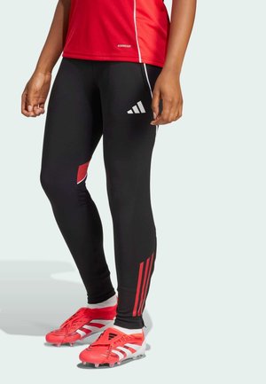 Leggings de sport noirs avec des accents rouges, dotés de trois bandes blanches sur le côté. Portés avec des chaussures de football rouges mettant en valeur des détails blancs.