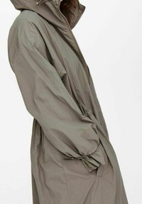 Luftigt, långt khaki regnjacka med huva, elastiska ärmar och framdraget blixtlås, bärs av en person som står i profil mot en enkel bakgrund.