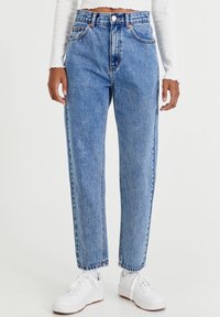 Vaaleansinisiä korkeavyötäröisiä denim-farkkuja suoran lahkeen istuvuudella, joissa on etutaskut ja oranssit ompeleet, yhdistettynä valkoisiin tennareihin.