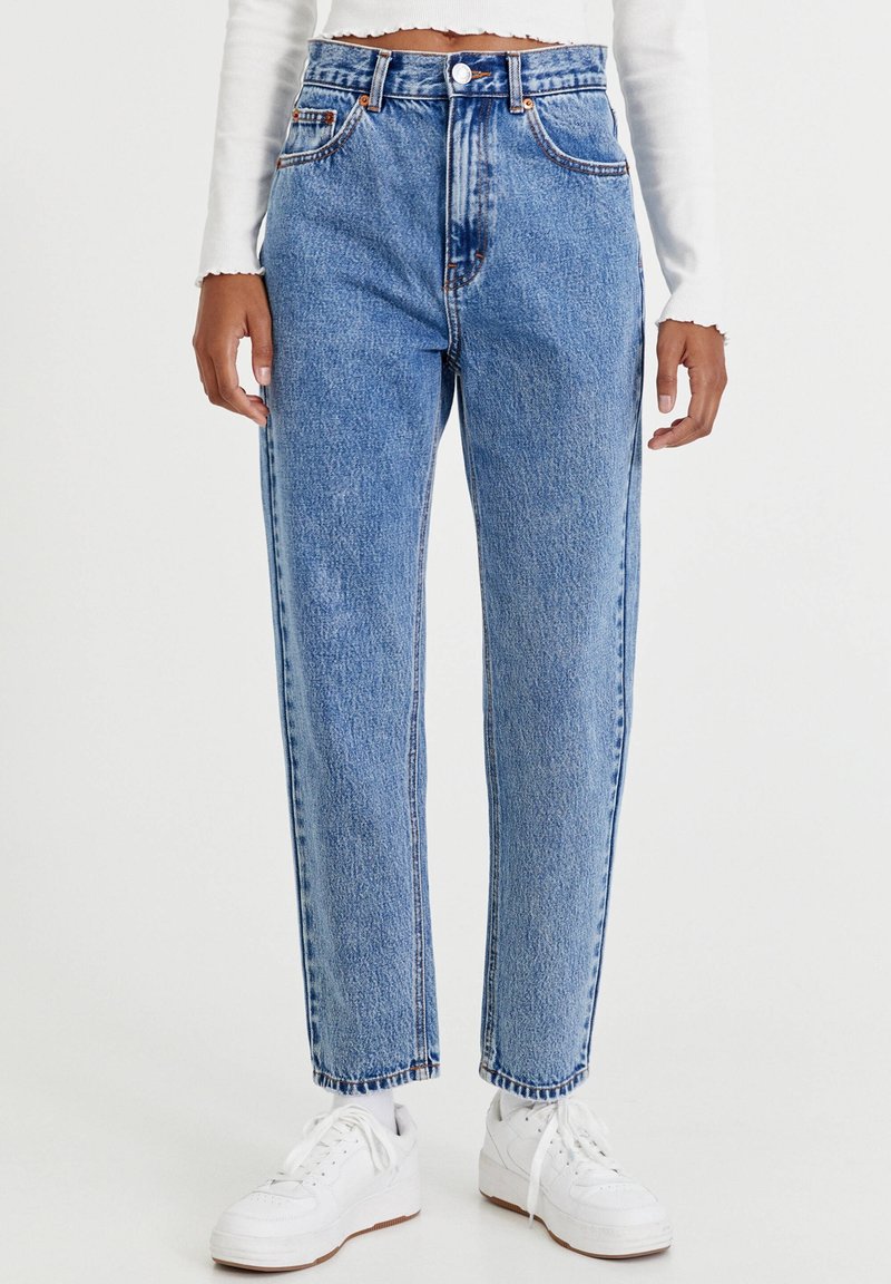 Vaaleansinisiä korkeavyötäröisiä denim-farkkuja suoran lahkeen istuvuudella, joissa on etutaskut ja oranssit ompeleet, yhdistettynä valkoisiin tennareihin.