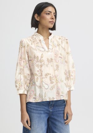Kvinde med kort mørkt hår iført en lyserød blomsterprint paisley-bluse med 3/4 ærmer og blå jeans, set fra siden mod en ensfarvet baggrund.