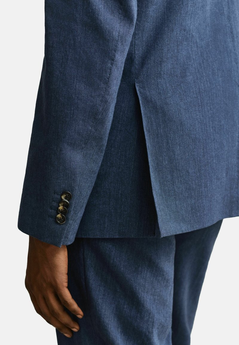 Primer plano de una mano de piel oscura junto a la manga de una chaqueta de traje azul marino que presenta cuatro botones oscuros y una abertura en la parte trasera.